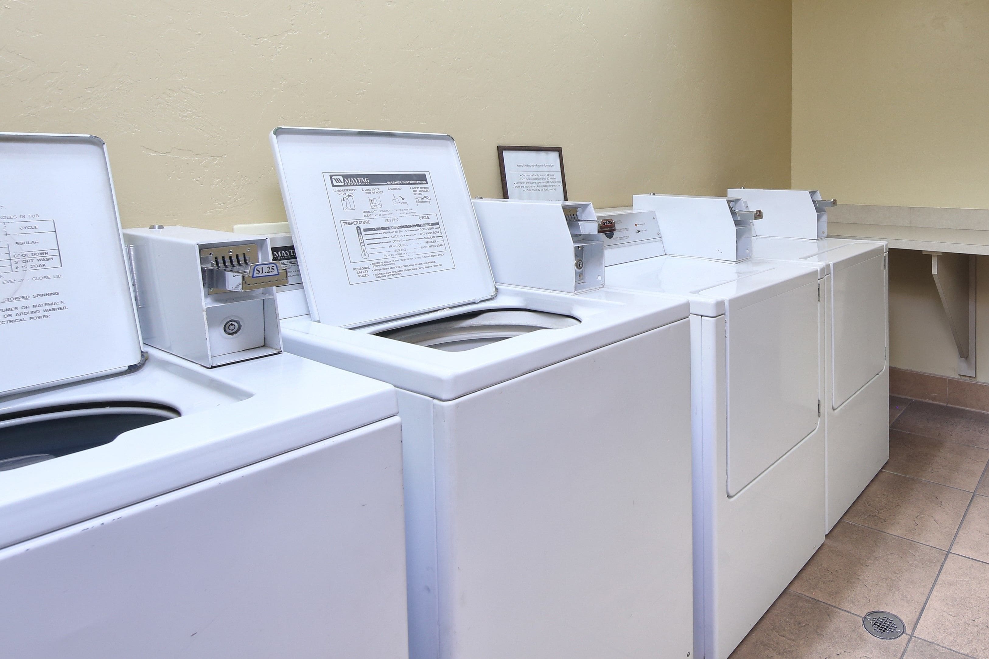 A row of white front-loading washers in a laundry room with beige walls and tiled flooring.