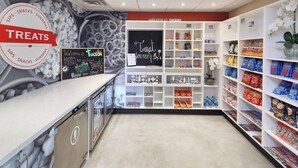 A clean, well-organized snack and sundries section with a variety of packaged snacks and beverages, a chalkboard sign welcoming visitors, and a decorative wall with a circular sign indicating the treats available. - Hampton Inn & Suites Tucson-Mall (Tucson)