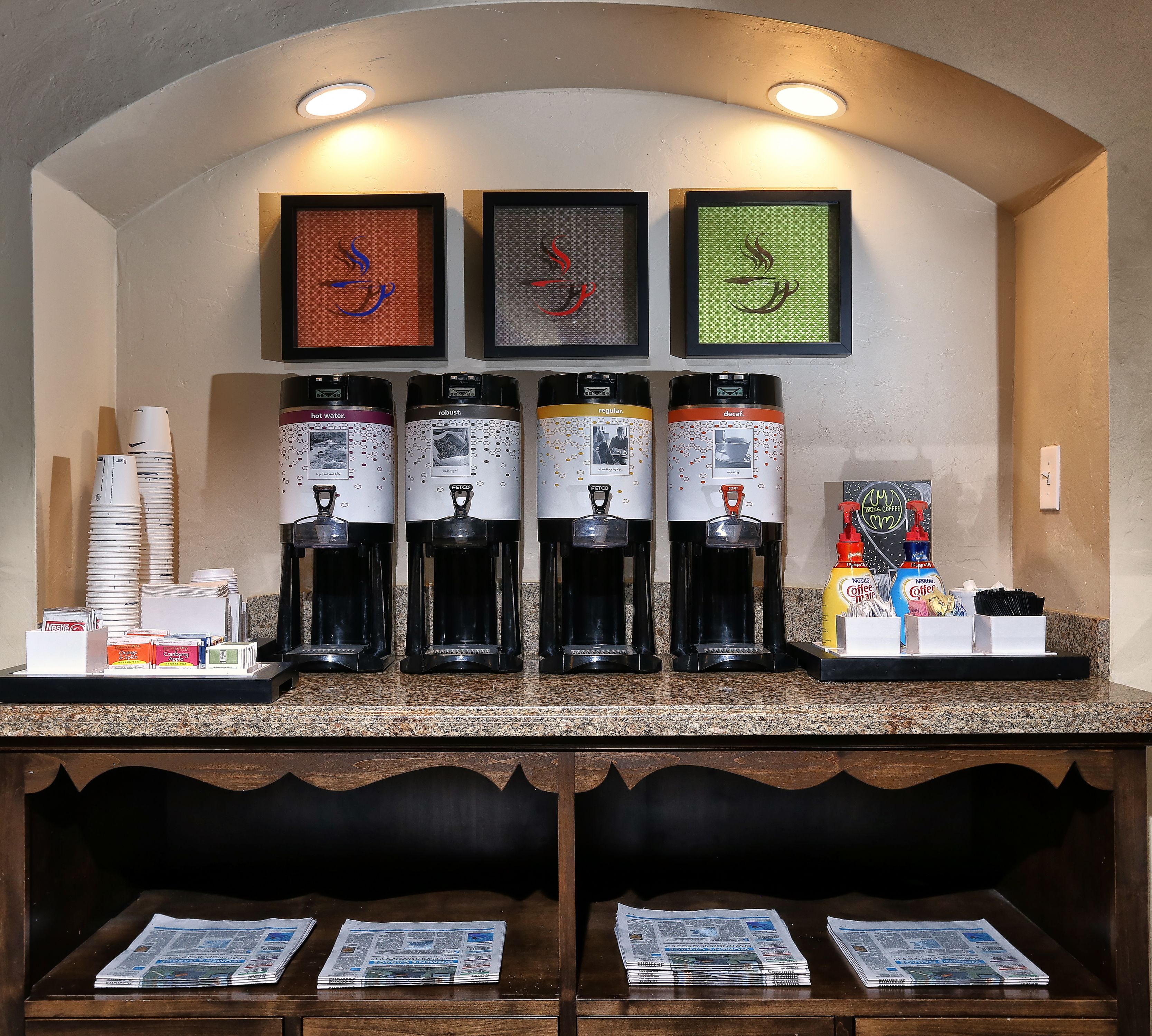 Una estación de café con varios dispensadores etiquetados para agua caliente, café intenso, café normal y café descafeinado. Hay pilas de vasos desechables y una variedad de cápsulas de café.