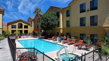 A pool area with lounge chairs and a hot tub, surrounded by a fence and a building with multiple windows.