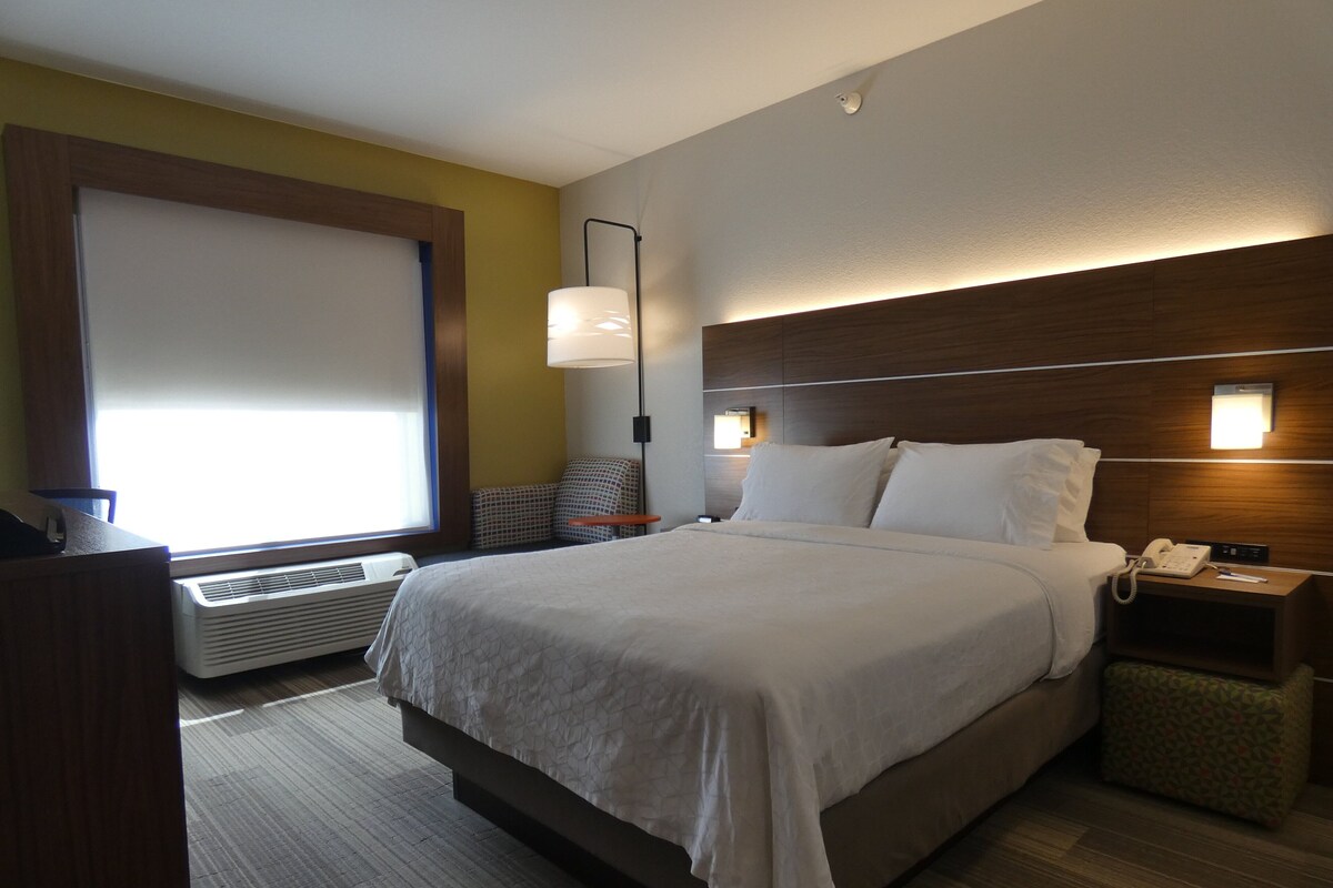 A hotel room with a bed, bedside table, lamp, and a window with blinds.