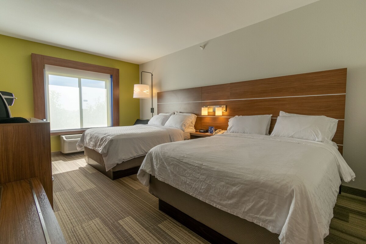 A hotel room with two beds, a desk, a lamp, and a window.