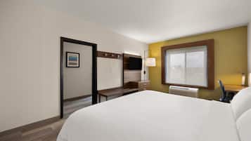 A hotel room with a large bed, a desk with a flat-screen TV, a window with blinds, and a door with a sign.