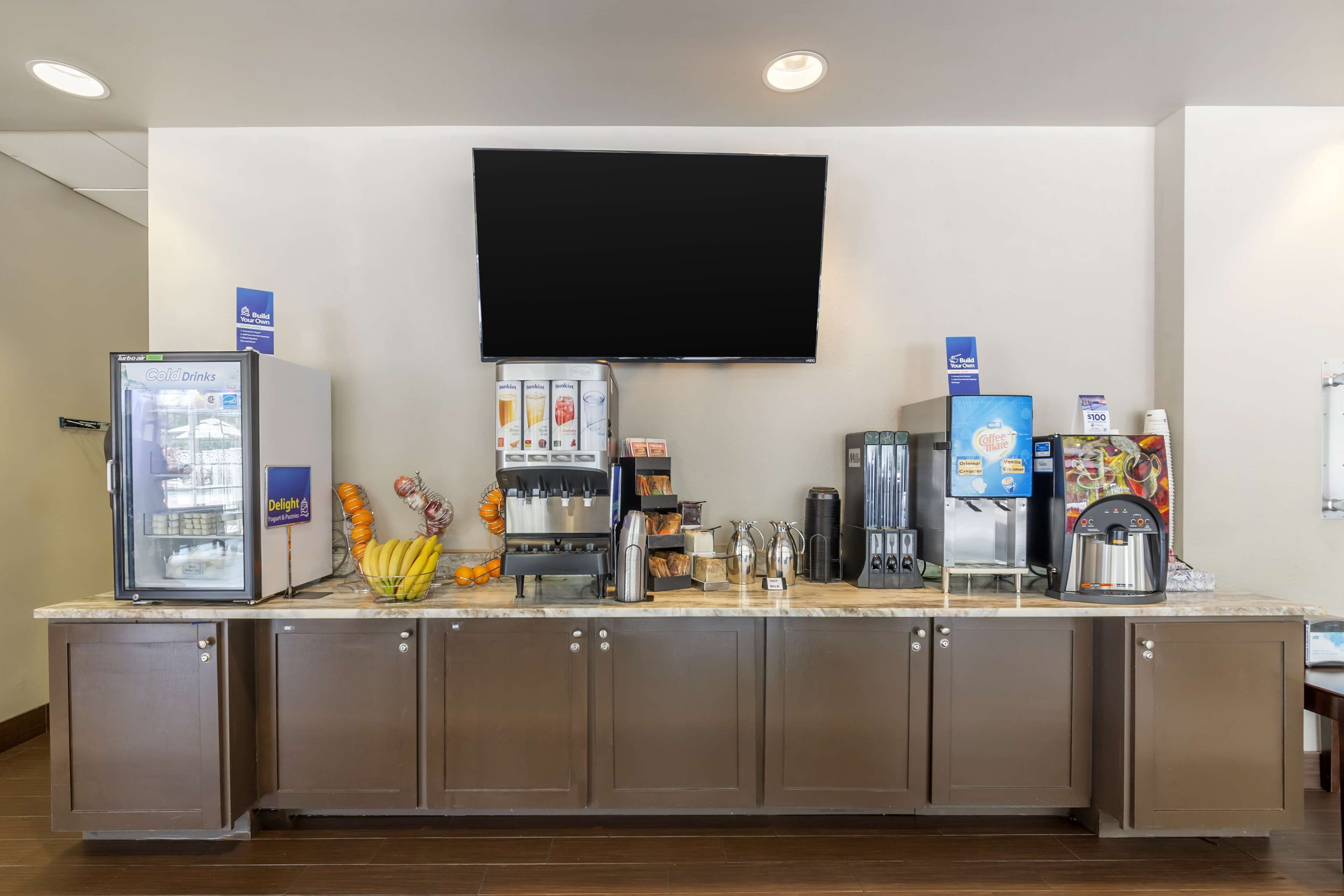 Un angolo colazione self-service con un frigorifero, macchine da caffè e un televisore a schermo piatto montato a parete.