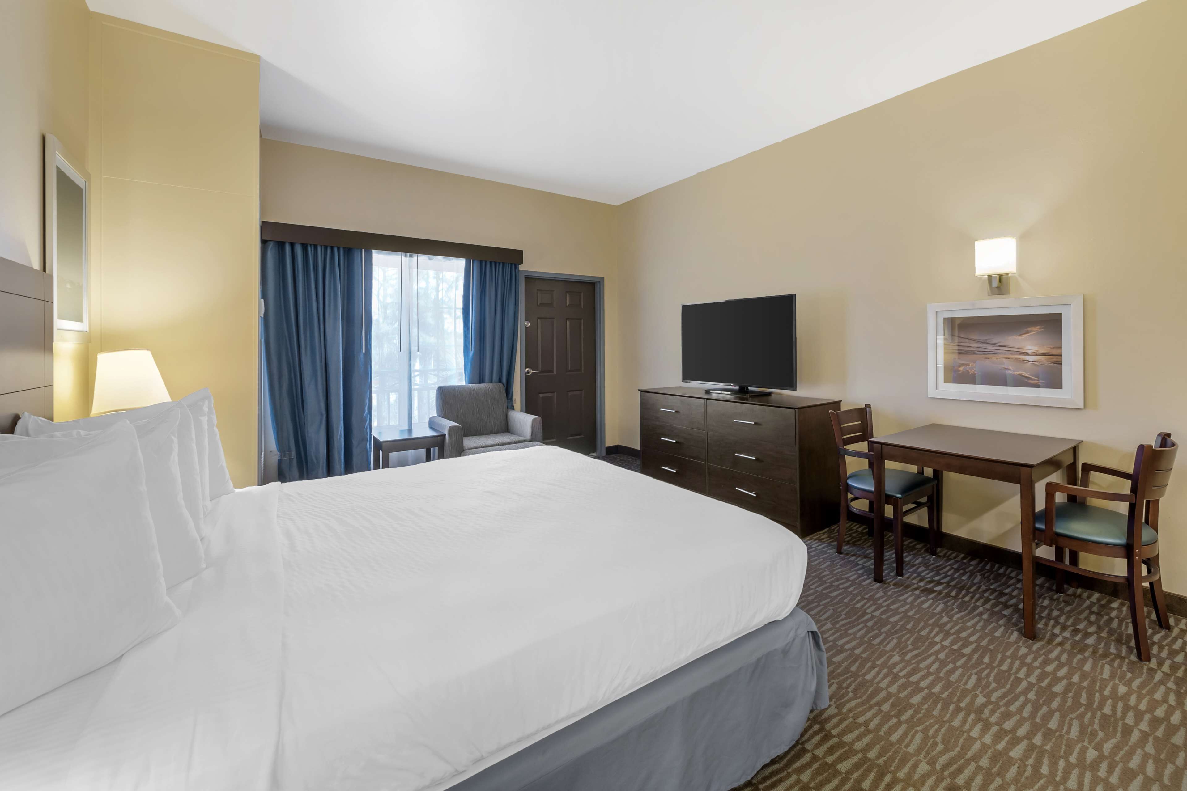 A hotel room with a large bed, a desk with two chairs, a television, a dresser, and a window with curtains.
