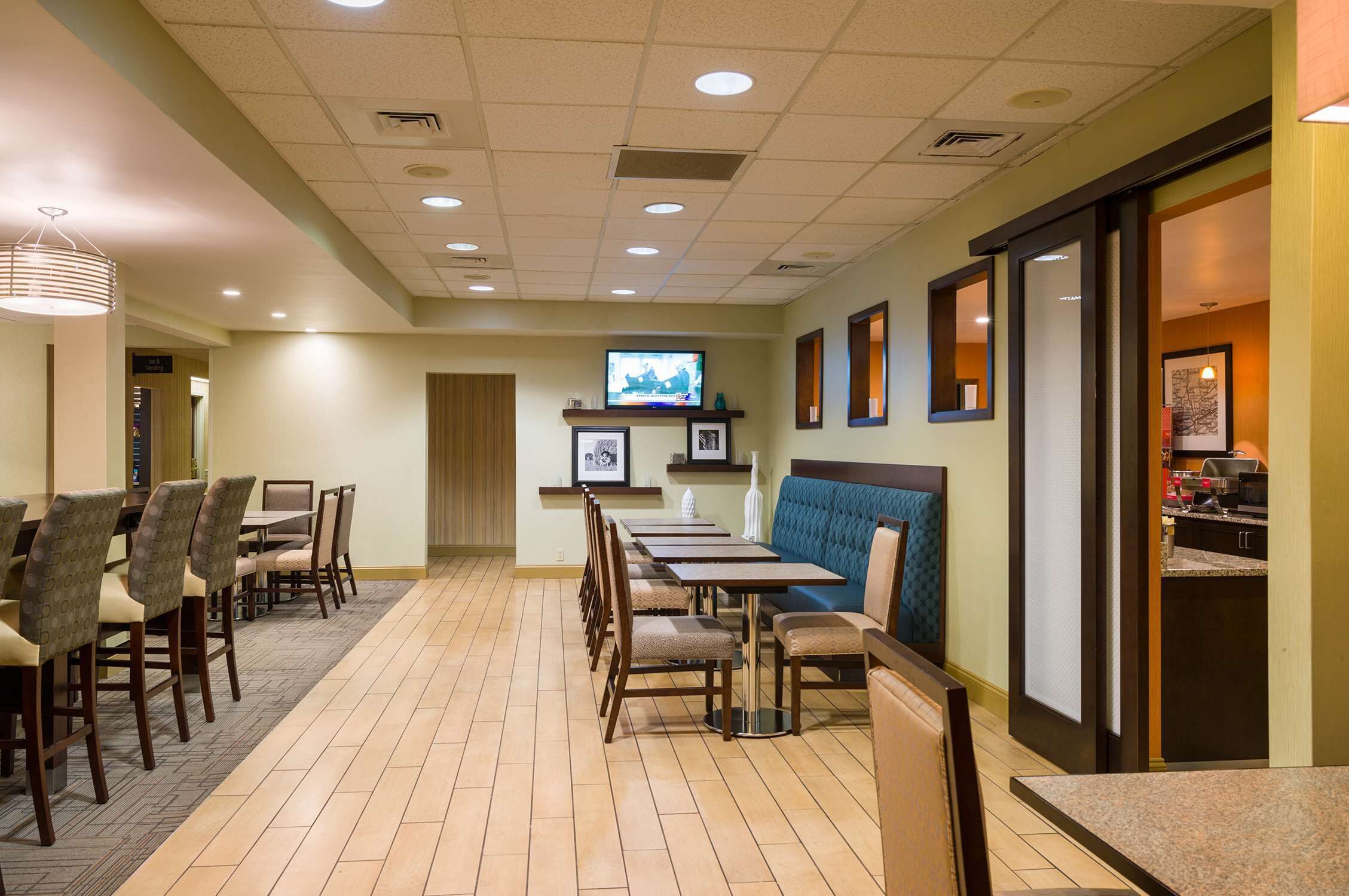 A hotel dining area with multiple tables and chairs, a television mounted on the wall, and a kitchen area visible through an open door.