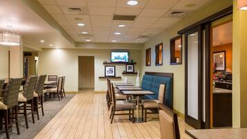 A hotel dining area with multiple tables and chairs, a television mounted on the wall, and a kitchen area visible through an open door.
