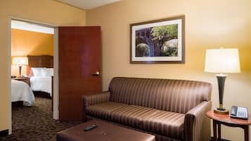 Une chambre d’hôtel avec un canapé à rayures, une petite table avec une lampe, une télécommande posée sur un pouf et un tableau encadré accroché au mur.
