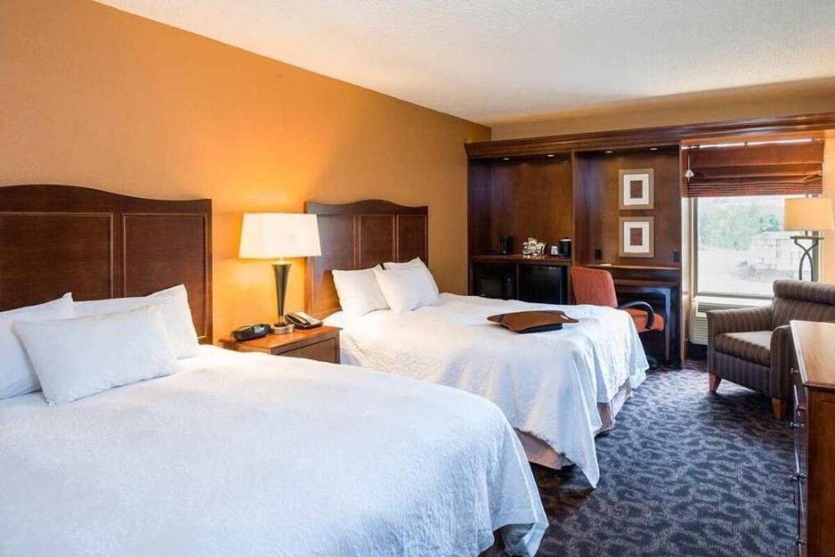 A hotel room with two beds, a desk, a chair, a lamp, and a window.