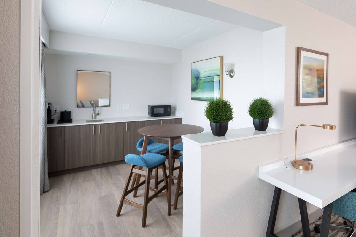 Una cocina moderna con armarios de madera, un microondas y una zona de comedor con sillas azules. Hay dos plantas en macetas sobre la encimera y un cuadro en la pared.