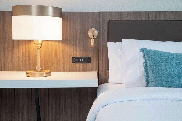 A neatly made bed with white and blue pillows, a blue throw blanket, a bedside lamp, and a wall-mounted light fixture.