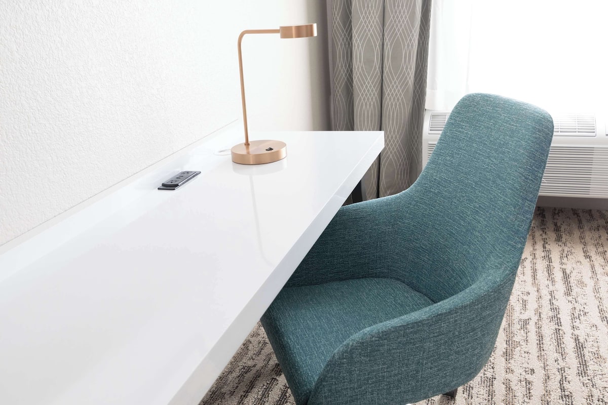 Una habitación de hotel moderna con un escritorio blanco, una silla color turquesa, una lámpara de pie y una ventana con cortinas.