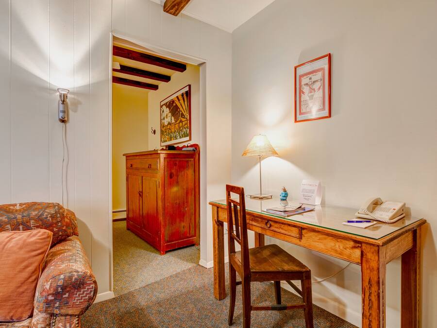 A small room with a wooden desk, a chair, a lamp, and a telephone. There is a couch with a patterned cushion and a door leading to another room.