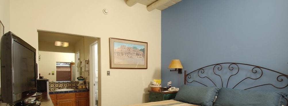 A bedroom with a bed, a nightstand, a lamp, a television, and a kitchen visible through an open door.