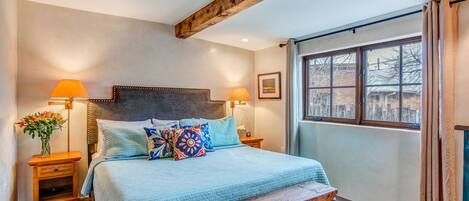 A bedroom with a large bed, two bedside tables, a bench, and a window with curtains.