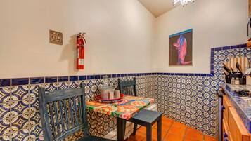 Una cocina con salpicadero de azulejos, un extintor de incendios en la pared, una mesa con mantel colorido y una silla.