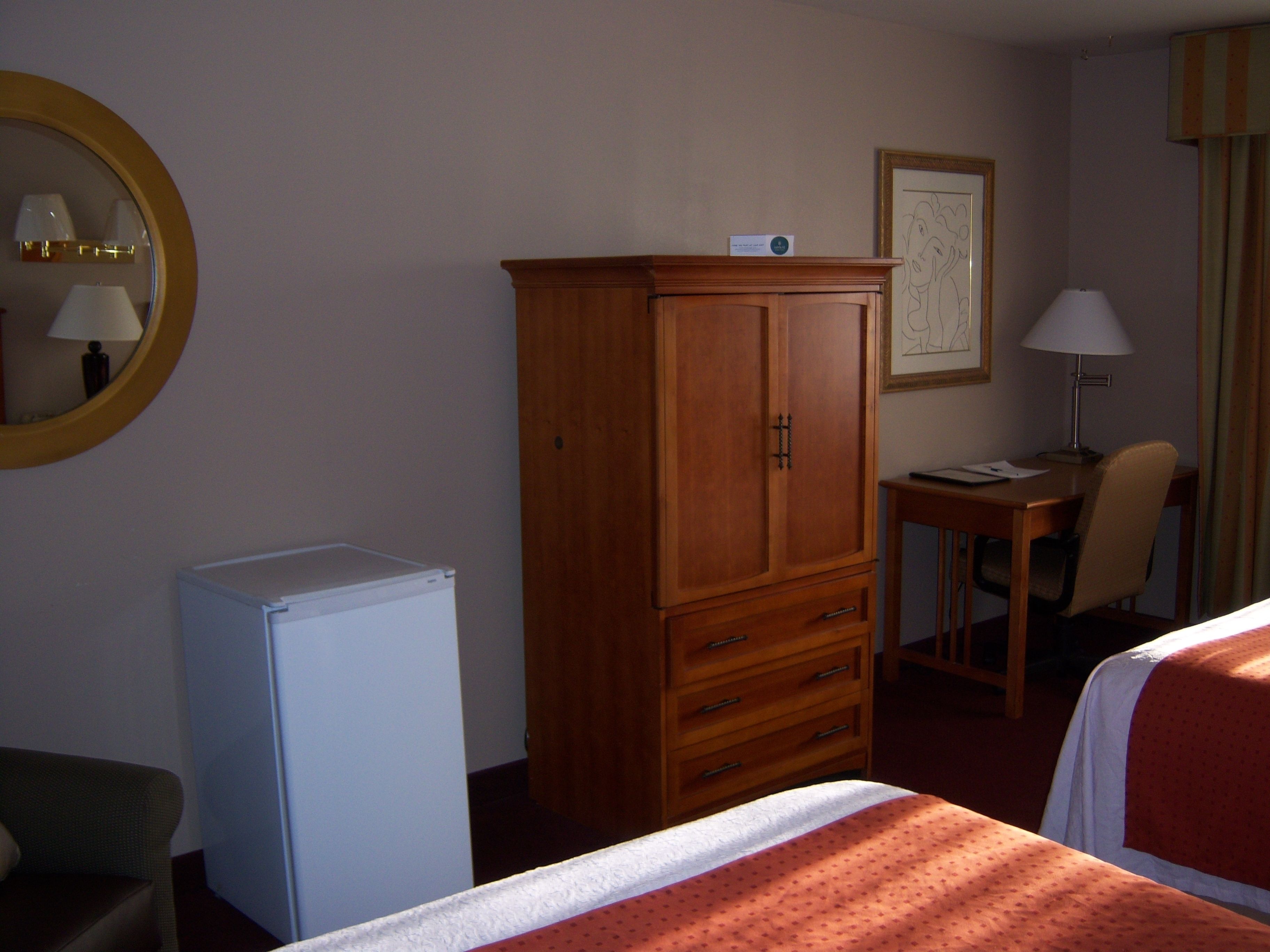 A hotel room with a wooden wardrobe, a desk with a chair, a bed, and a small white refrigerator.