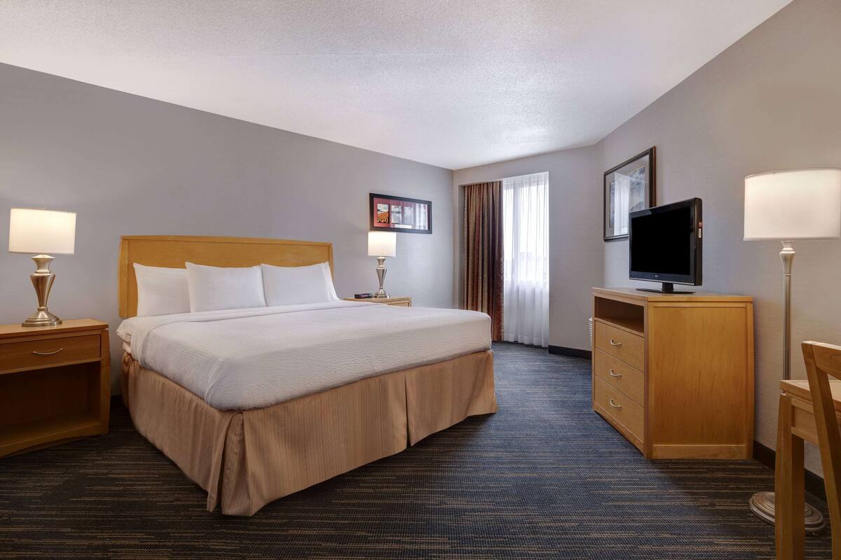 Une chambre d’hôtel comprenant un lit, deux tables de chevet avec des lampes, une télévision, une commode en bois et un sol recouvert de moquette.