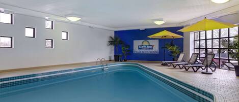 An indoor swimming pool with a blue tiled floor, a pool ladder, and lounge chairs with umbrellas.