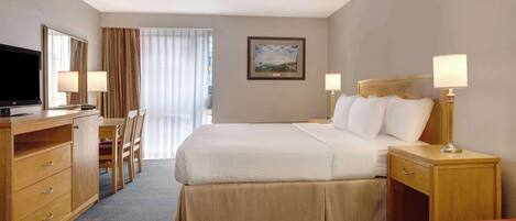 A hotel room with a bed, a desk with a TV, a chair, a nightstand with lamps, and a window with curtains.