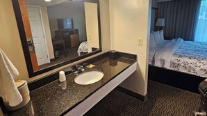 Ett hotellbadrum med en stor spegel, ett handfat och en säng som syns genom en öppen dörr.