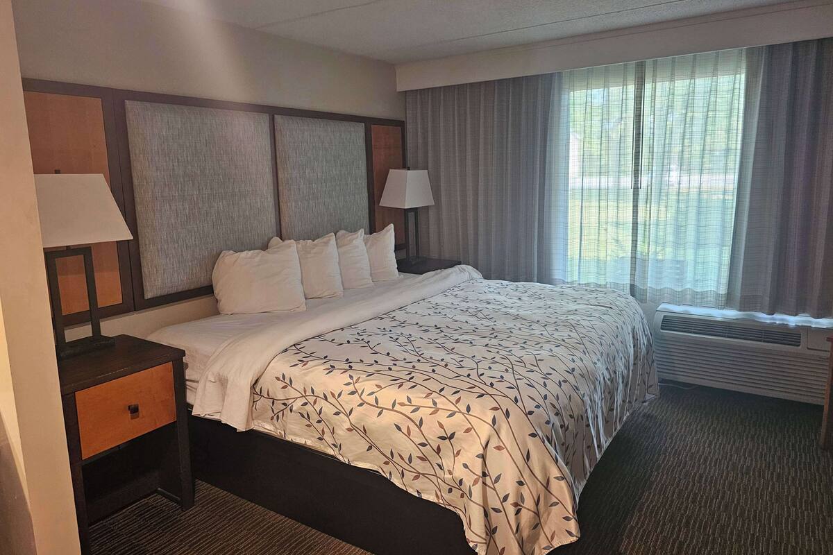 A hotel room with a large bed, two bedside tables with lamps, a window with curtains, and a wall-mounted air conditioning unit.