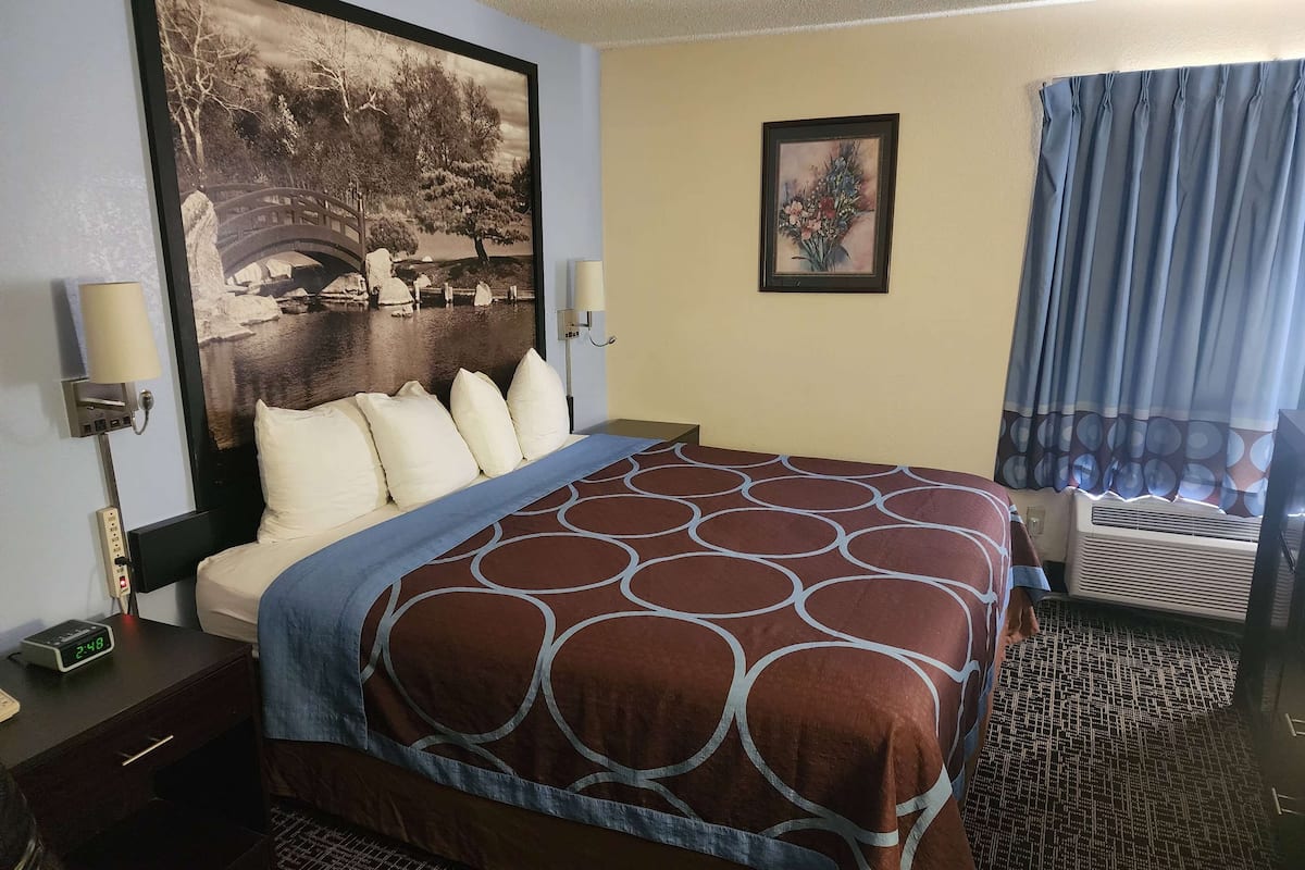 A hotel room with a bed, a nightstand, a lamp, a wall-mounted air conditioner, and a window with curtains.
