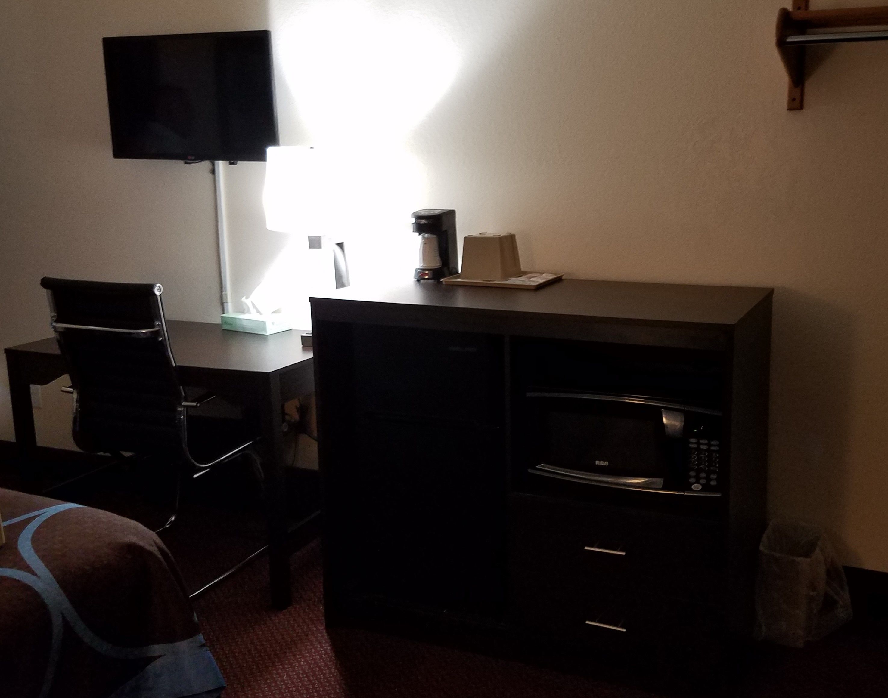 A small, dimly lit room with a desk, a chair, a microwave, and a television mounted on the wall.