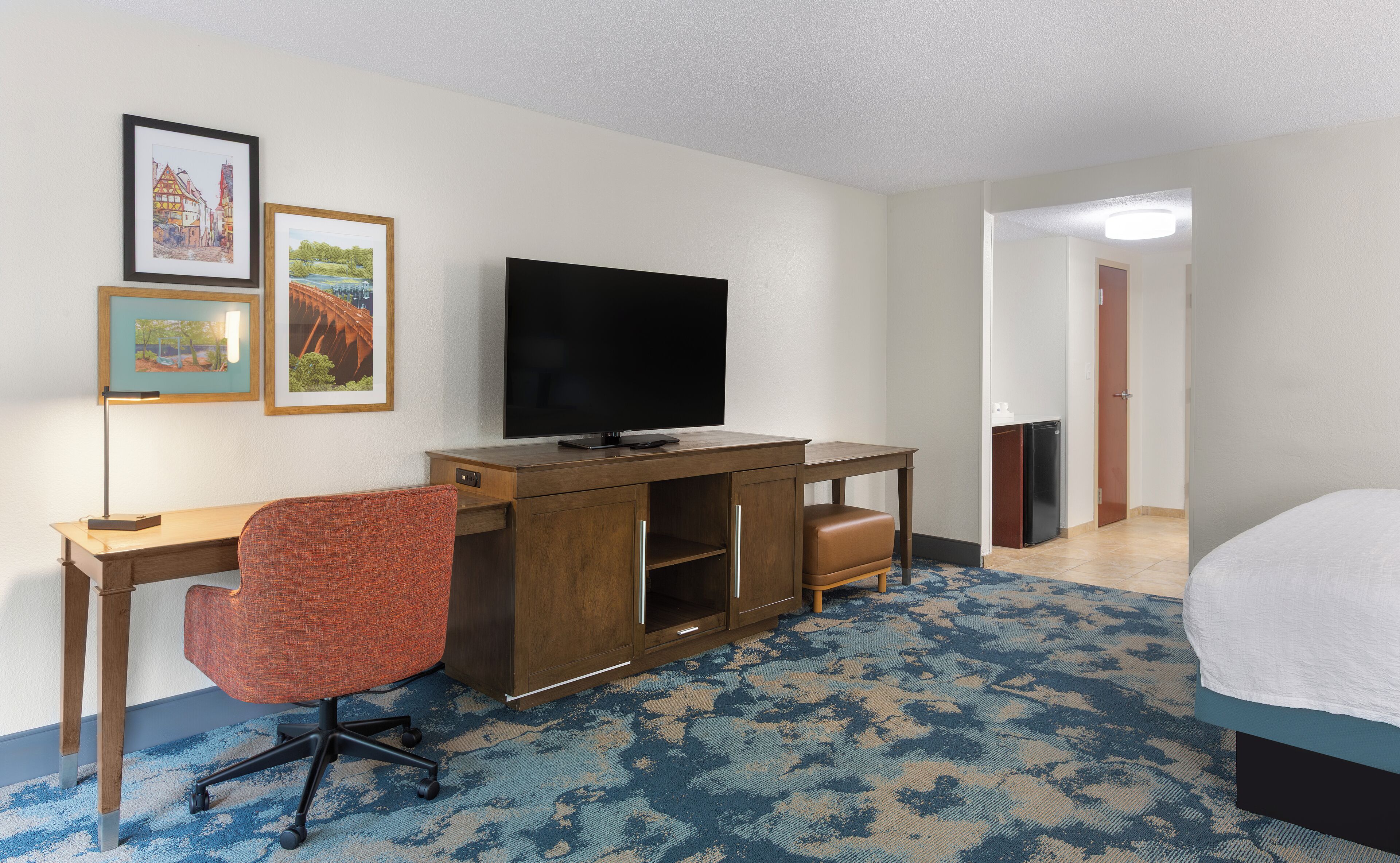 A hotel room with a bed, a desk with a TV, a chair, and two framed pictures on the wall.