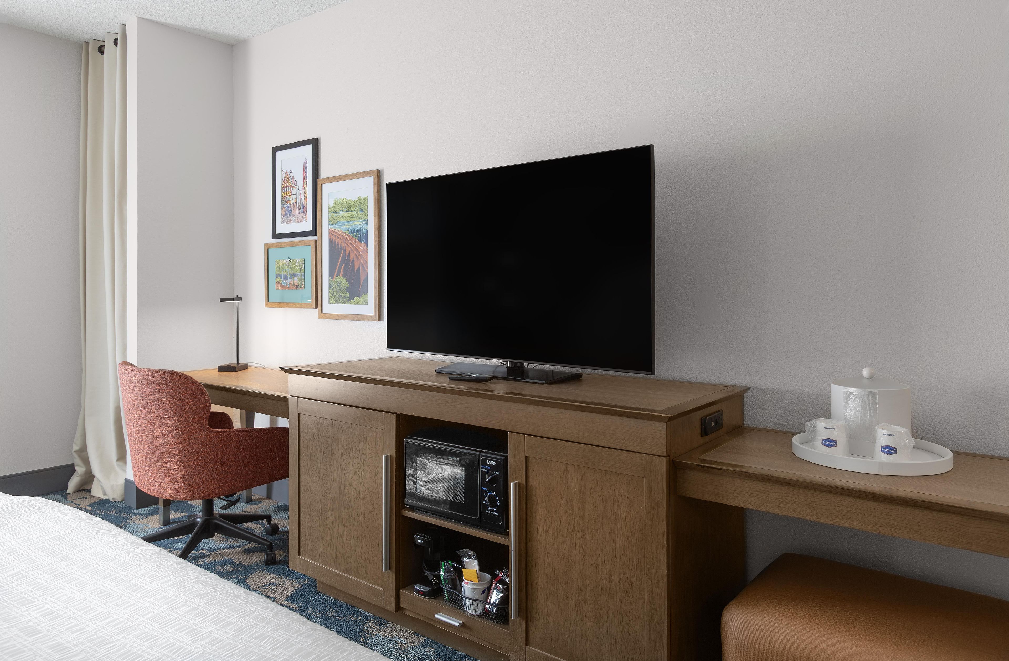 A hotel room with a bed, a desk with a flat-screen TV, a red chair, and a coffee machine.