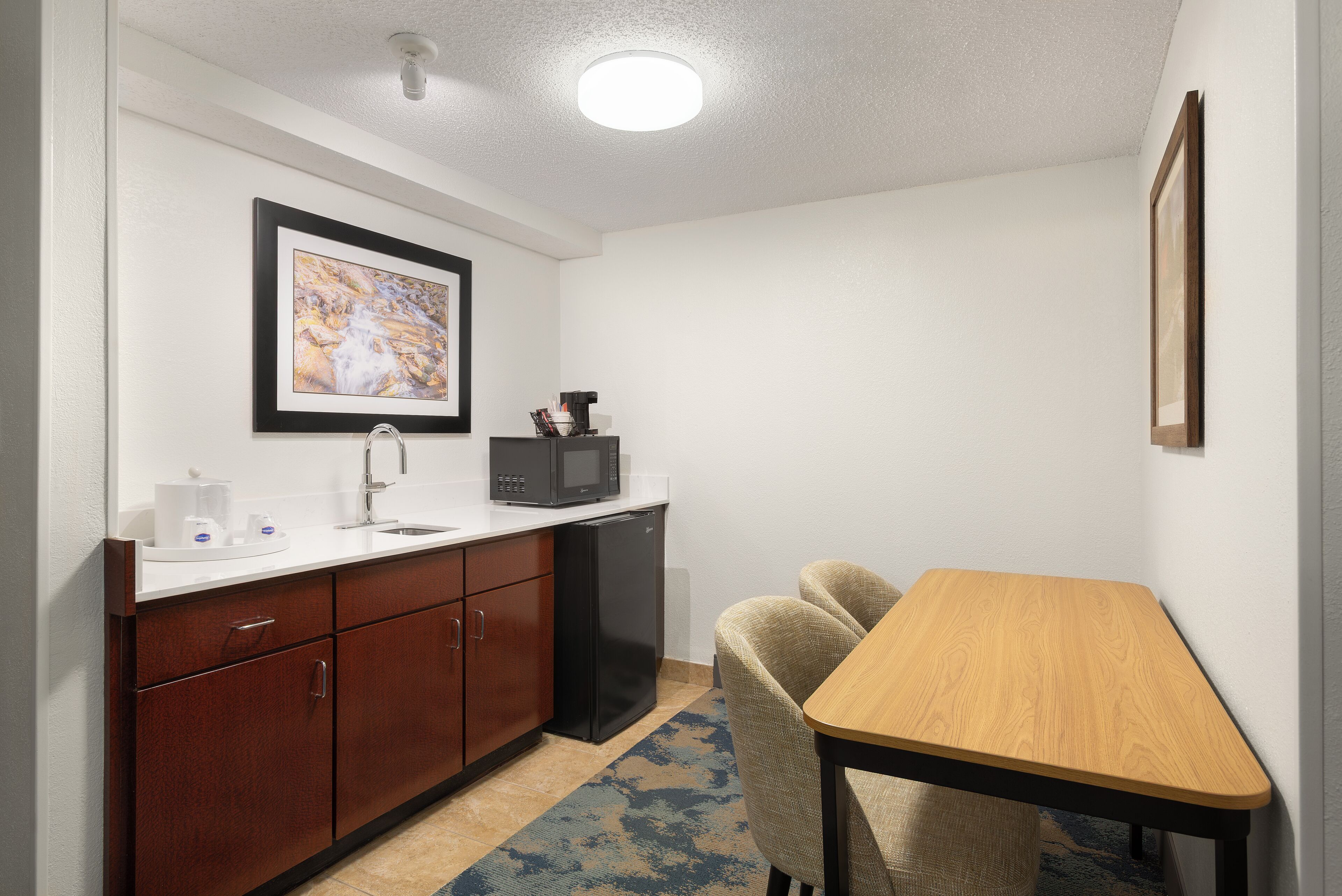 A compact kitchen with a microwave, coffee maker, and sink. There is a framed picture on the wall and a small table with chairs.