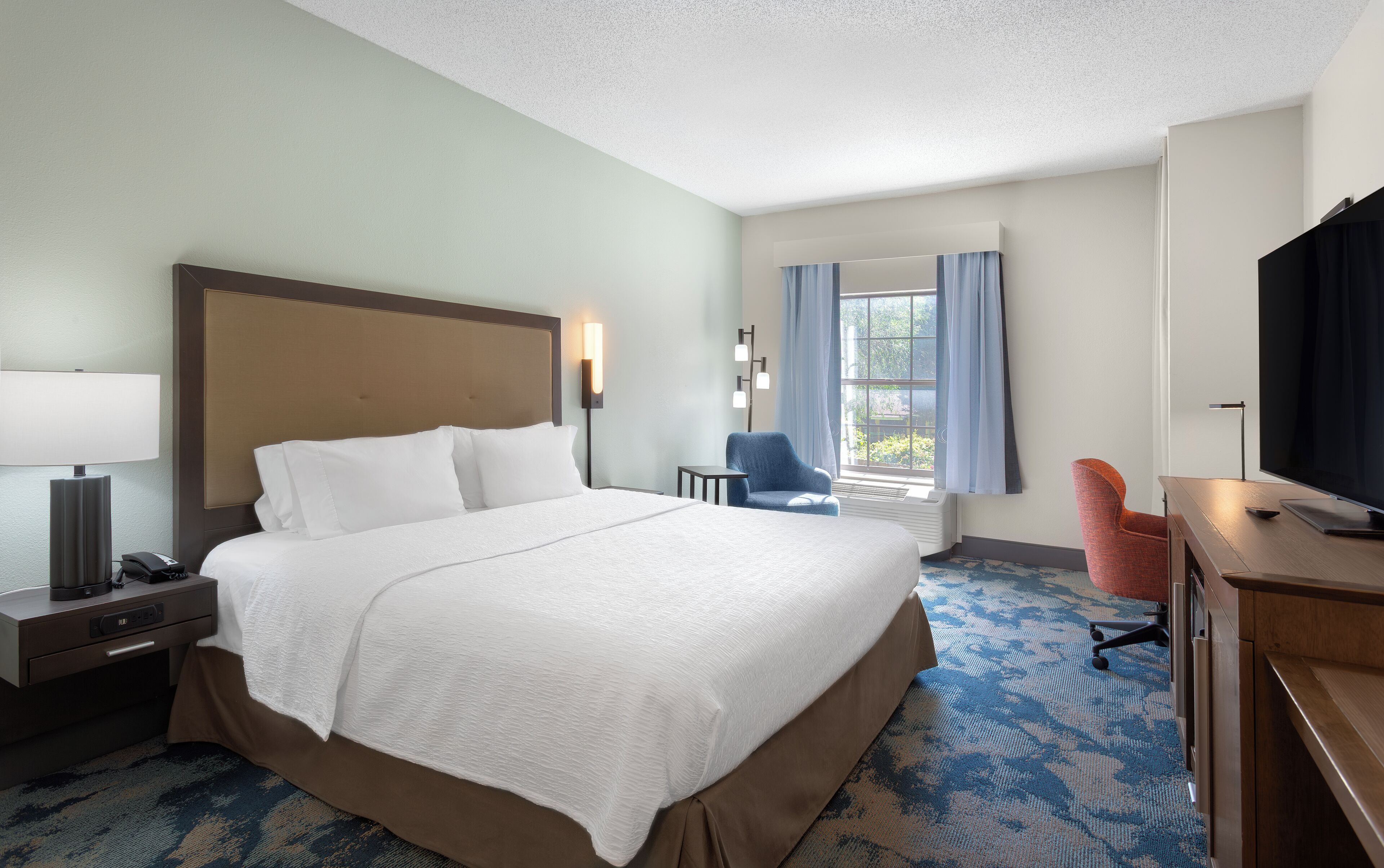 A hotel room with a large bed, bedside table with a lamp, a chair, a TV, and a window with curtains.