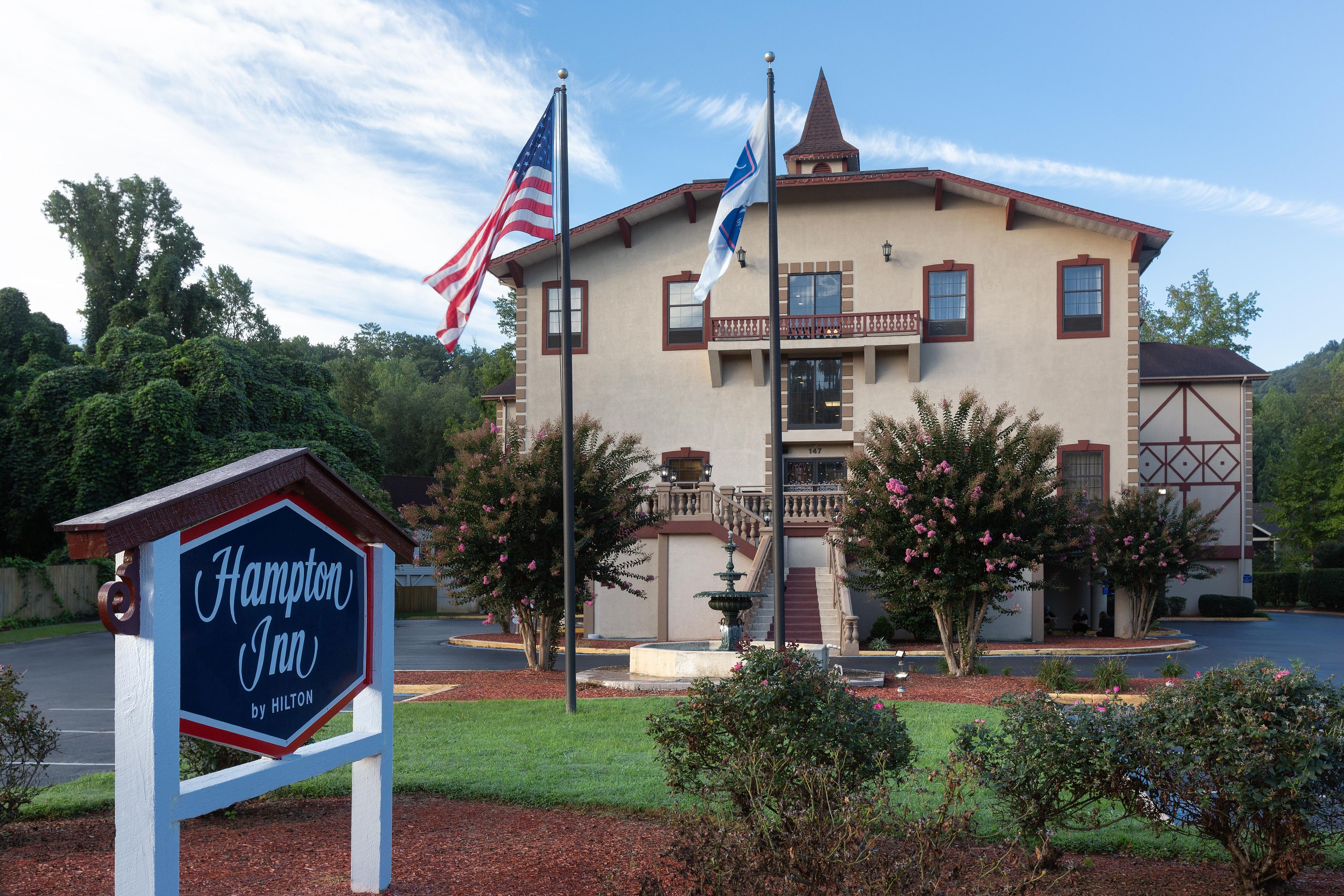 Ein Hotel mit Hinweisschild, das es als Hampton Inn by Hilton kennzeichnet, mit einem zweistöckigen Gebäude, einem Balkon, einer Fahnenstange mit der US-amerikanischen Flagge und einem gepflegten Garten.