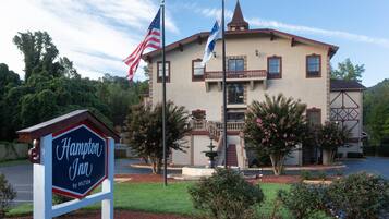 A hotel with a sign indicating it's a Hampton Inn by Hilton, featuring a two-story building with a balcony, a flagpole with the American flag, and a well-maintained garden.