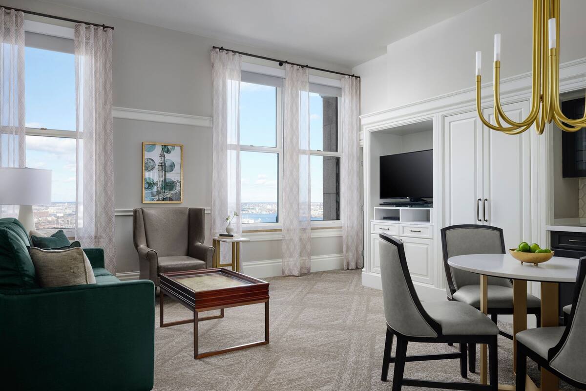 Un salon moderne avec un canapé vert, une table basse et un coin repas comprenant une table et des chaises. On aperçoit un téléviseur au fond et, grâce à de grandes fenêtres, on a une vue sur la ville.