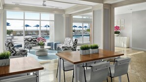 A spacious indoor area with a pool view, featuring a wooden table, white chairs, patterned sofas, and potted plants.
