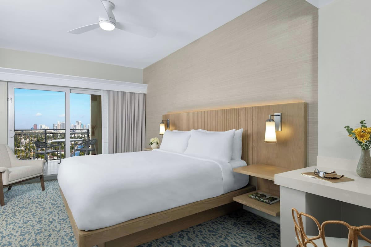 Een hotelkamer met een groot bed, een bureau, een stoel, een plafondventilator en uitzicht op de stad door grote ramen.