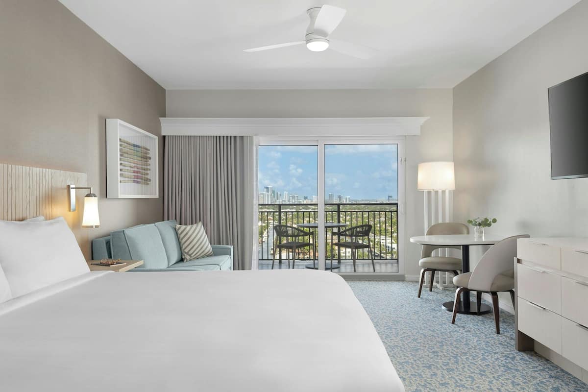 Een hotelkamer met een groot bed, een bank, een eettafel met stoelen, een balkon met uitzicht op de stad en een plafondventilator.