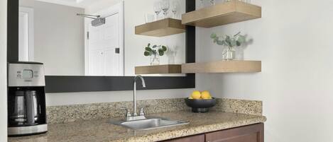 A kitchen with a granite countertop, a stainless steel sink, a coffee maker, wooden shelves with glasses, and a bowl of lemons.