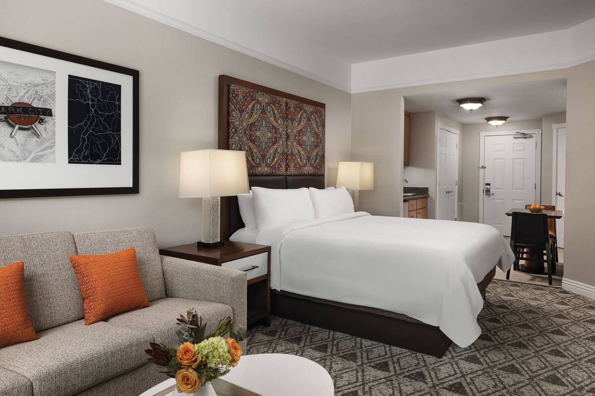 A hotel room with a bed, two bedside lamps, a sofa with orange cushions, a coffee table with flowers, and a framed cityscape artwork.