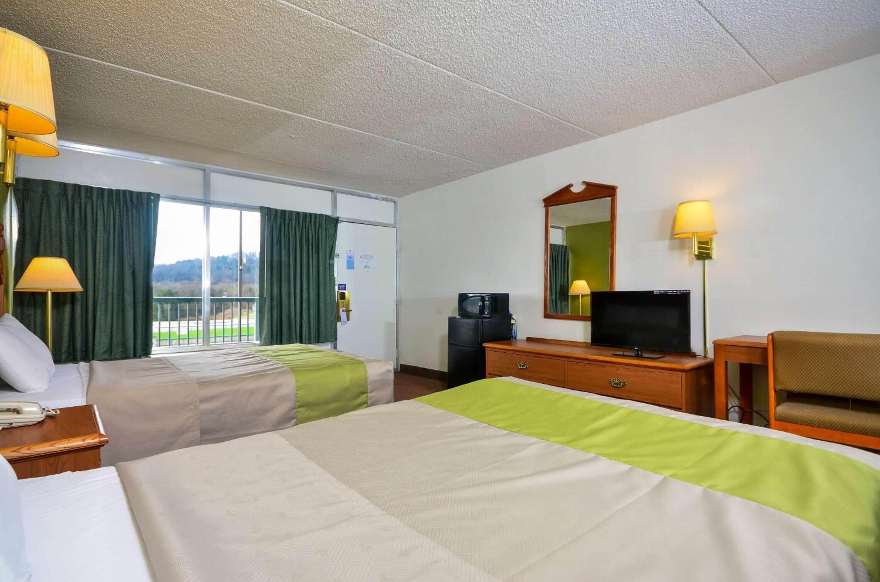 A hotel room with two beds, a television, a desk with a lamp, and a view of the outdoors.