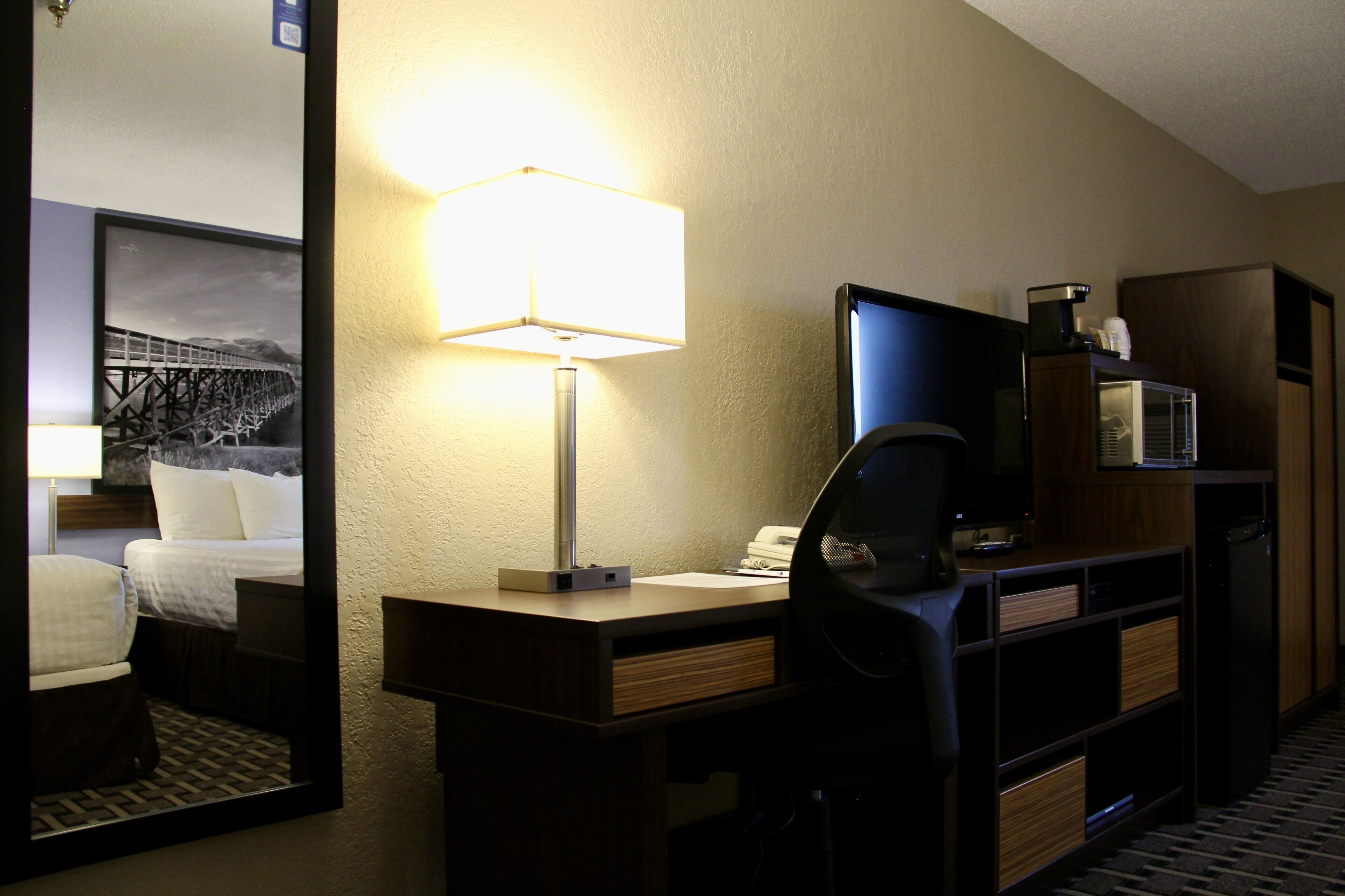 A hotel room with a desk, a lamp, a television, a mirror, a bed, and a wardrobe.