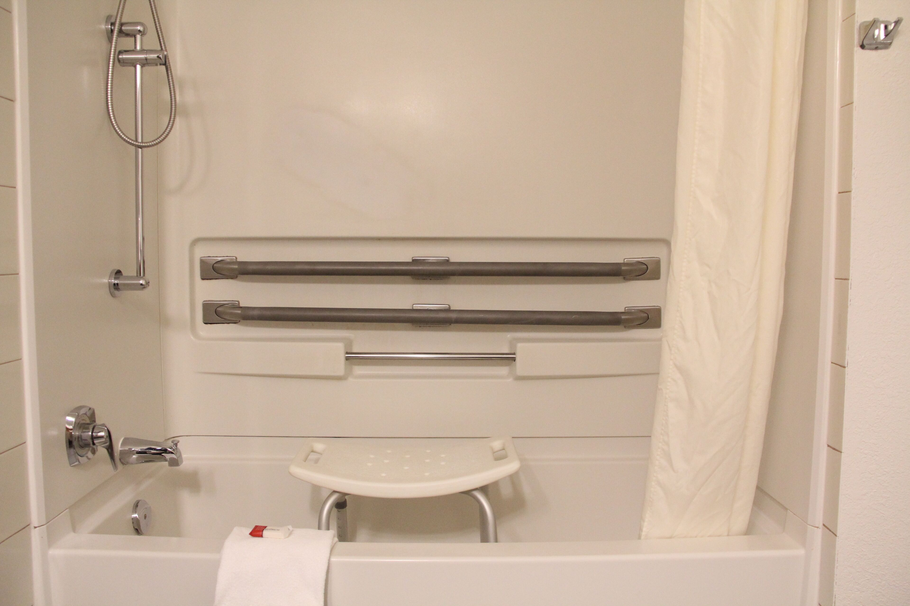 A bathroom with a bathtub, a shower, a grab bar, a small white stool, and a towel rack.