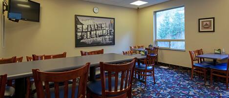 A conference room with a large table, multiple chairs, a mounted TV, a clock, and a window with a view of trees.
