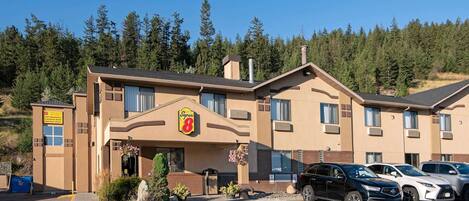 Un hotel con un logotipo destacado, un estacionamiento con varios coches y un telón de fondo natural de bosque.