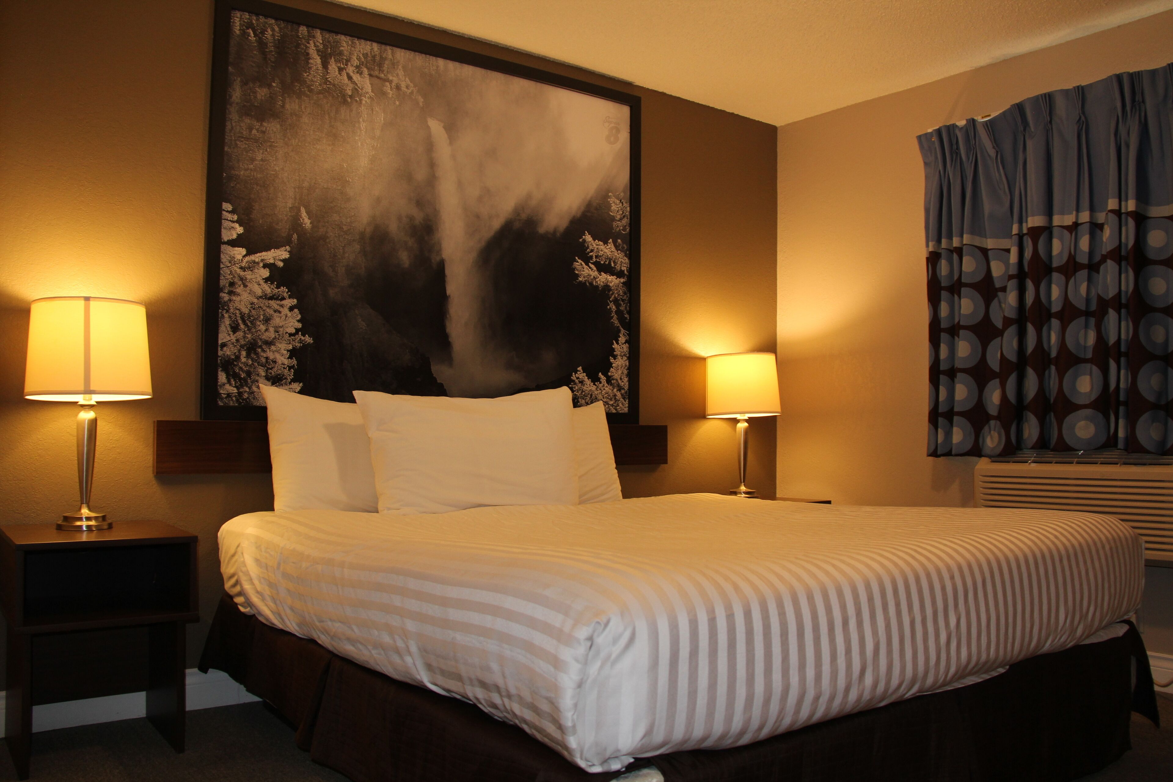A hotel room with a large bed, two bedside lamps, a large abstract painting on the wall, and a window with patterned curtains.
