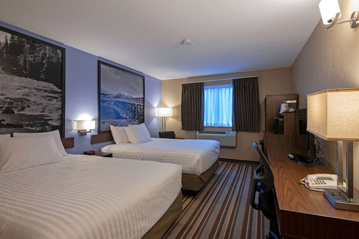 Quarto de hotel com duas camas, uma mesa de trabalho com telefone, uma luminária, uma TV e uma janela com cortinas.