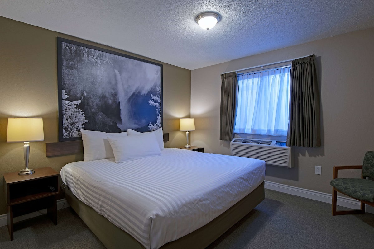 Quarto de hotel com uma cama grande, duas mesas de cabeceira com abajures, um quadro grande na parede, uma janela com cortinas e uma cadeira.