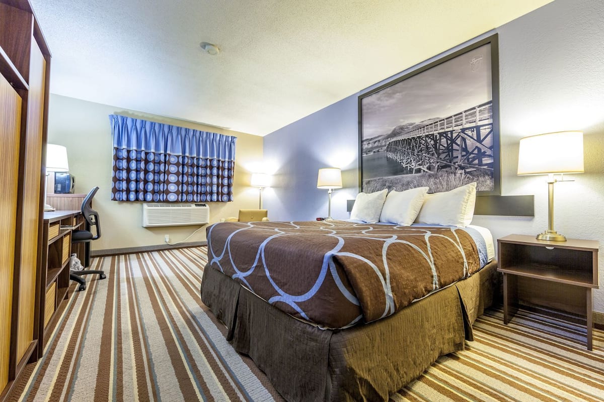 Quarto de hotel com cama, escrivaninha, cadeira, luminárias e uma grande moldura na parede.
