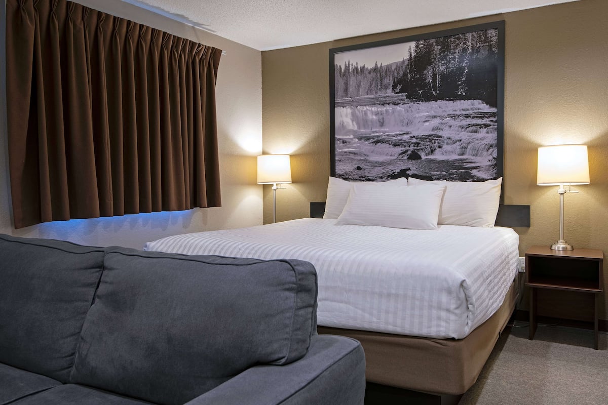 Quarto de hotel com uma cama grande, duas luminárias de cabeceira, um sofá cinza e uma grande moldura com uma imagem de uma cascata na parede.